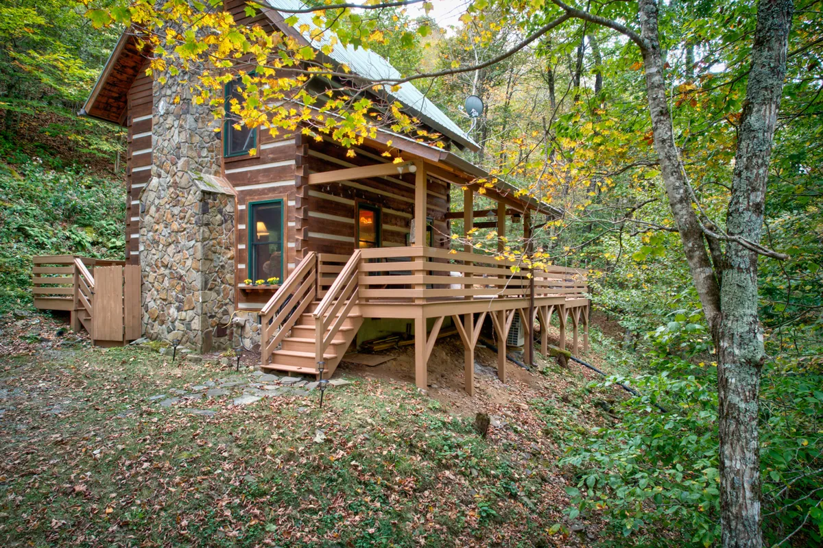 The image shows a charming log cabin set amidst a tranquil forest, with a sturdy stone chimney climbing up the side. A spacious deck with rustic railings invites guests to relax and soak in the surrounding natural beauty. Trees encircle the property, providing privacy and a sense of seclusion, while hints of sunlight filter through the branches, creating a warm, magical glow.