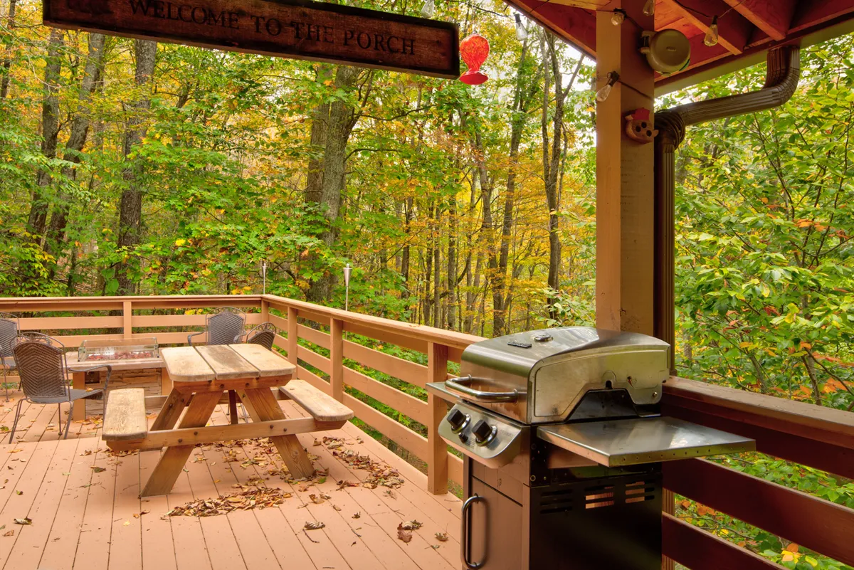 A spacious porch with a wooden picnic table and grill is framed by lush trees, bathed in warm evening light. String lights add a festive touch, creating an inviting atmosphere for outdoor dining, barbecues, or simply relaxing with loved ones after a day of local exploration.