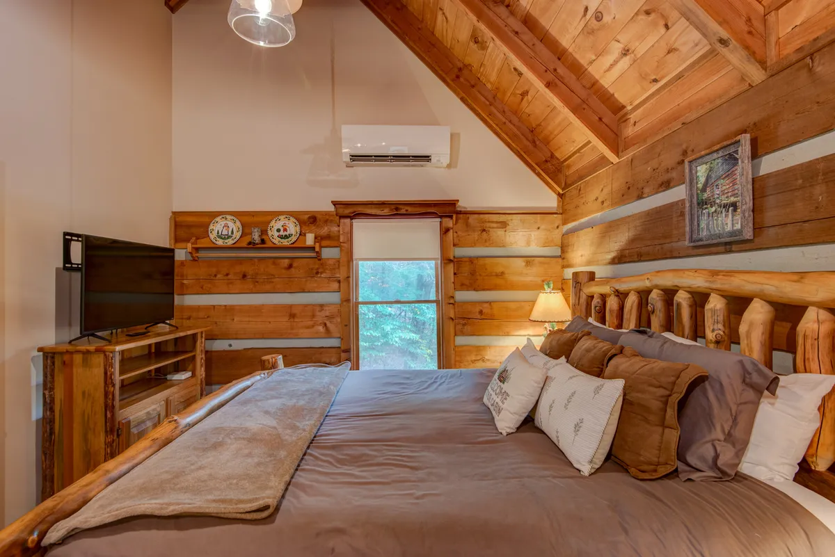 A cozy bedroom with a wooden bed frame, a television mounted opposite the bed, a window bringing in natural light and views of the outdoors, and decorative plates on the wall. A table lamp by the bedside adds a warm glow, and the arrangement feels both restful and inviting.