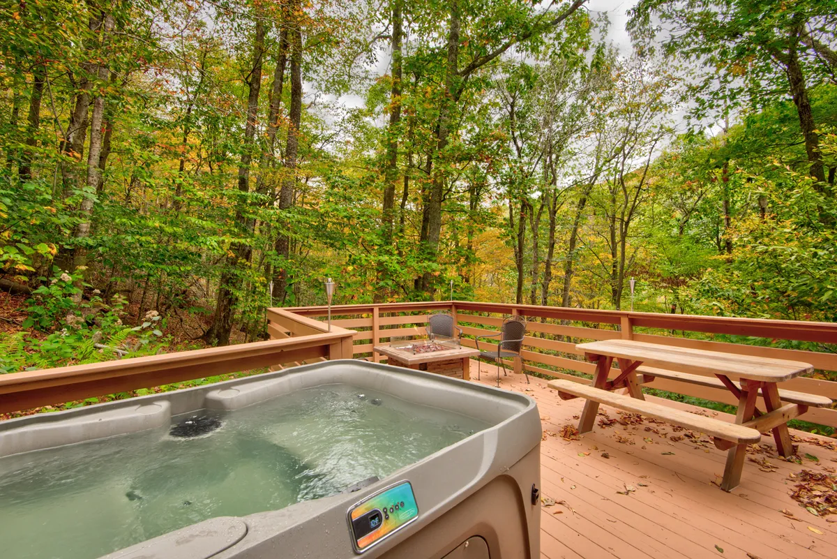 This image showcases a bubbling hot tub set on a wooden deck surrounded by tall trees, with comfortable outdoor seating and a picnic table also visible. The scene is bathed in serene sunset light, emphasizing the feeling of a tranquil forest retreat.