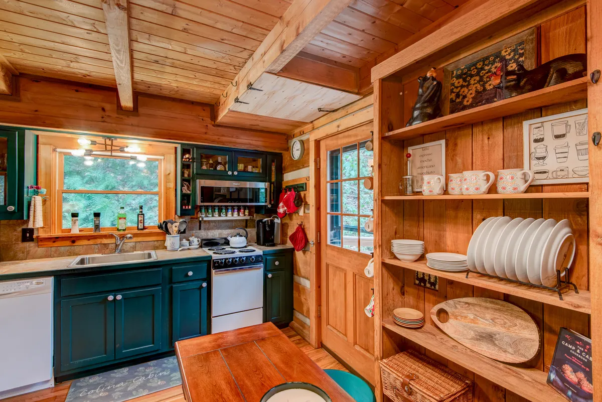 A spacious kitchen and dining area with a prominent wooden countertop, green cabinets, open wooden shelves, and a large dining table. The setting appears inviting and ready for both cooking and gathering around the table.