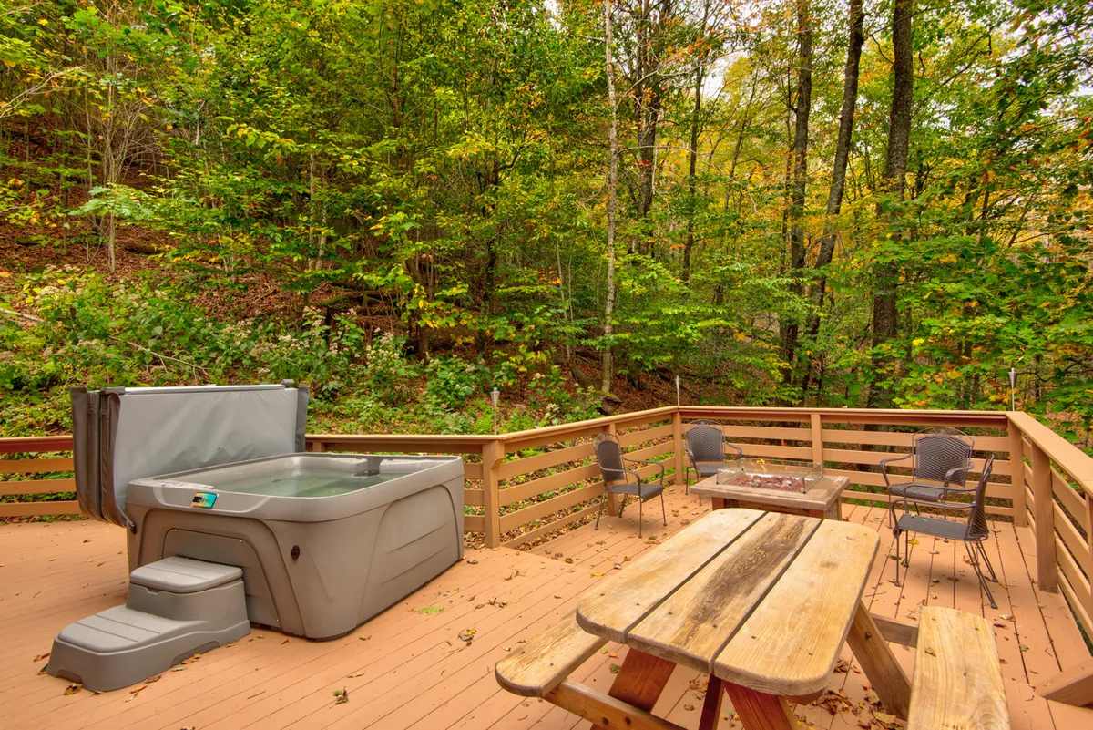 A spacious wooden deck with a bubbling hot tub, outdoor seating, and lush trees all around. The serene atmosphere is enhanced by the possibility of stargazing from the hot tub after a day of exploration, or socializing with friends and family.