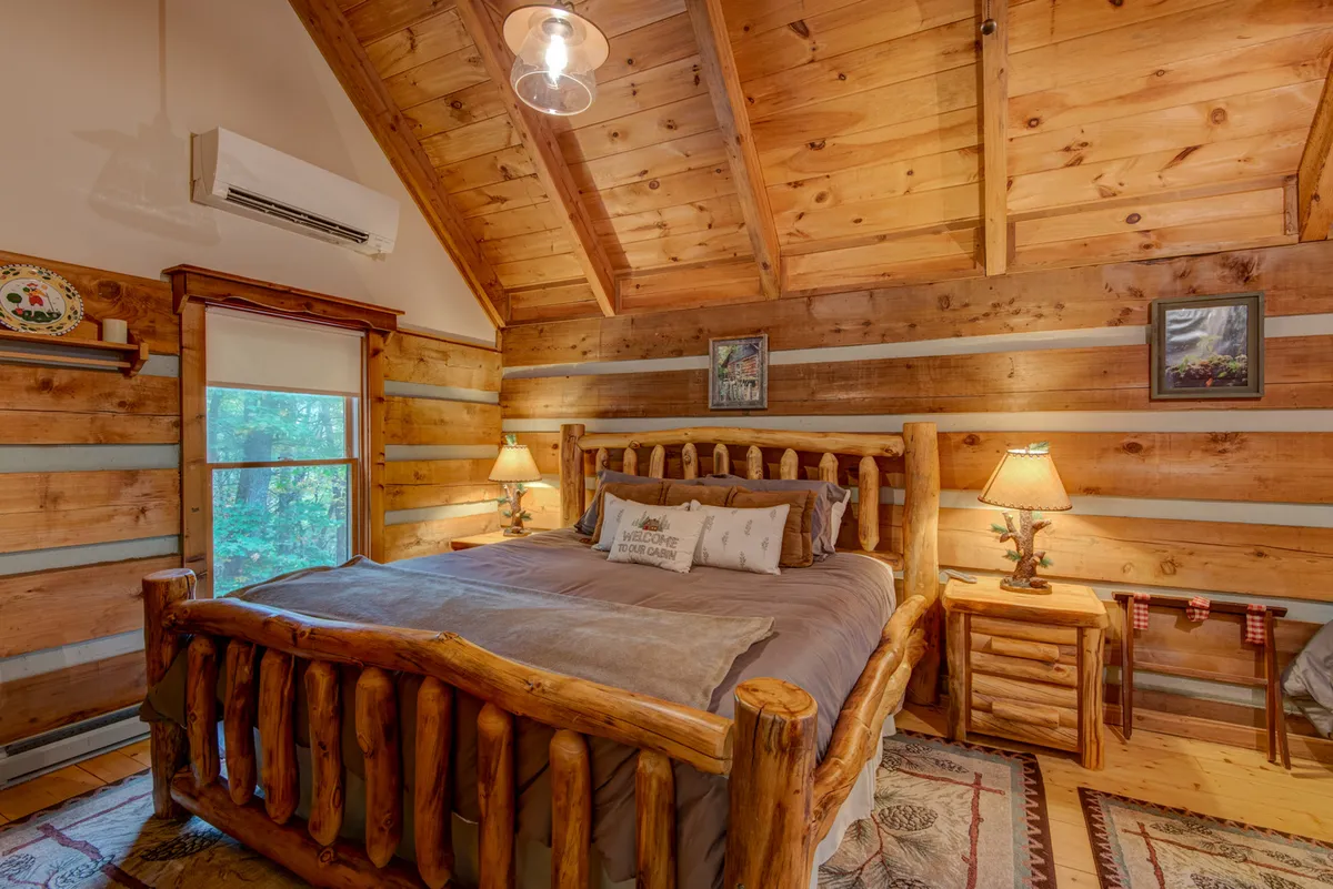 A log-framed bed with soft bedding is set against wooden walls, flanked by nightstands topped with lamps and decorative artwork above the headboard. The space exudes warmth and tranquility, ideal for relaxation or quiet moments.