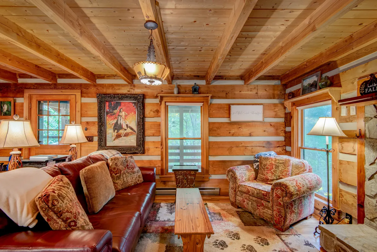A leather sofa anchors this warmly lit living area, joined by a floral armchair, wooden coffee table, framed artwork, and window views. The arrangement suggests relaxed conversations, shared drinks, and a welcoming space for movie nights or quiet moments alike.