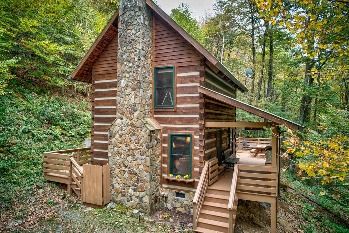 An inviting view of the log cabin's exterior, featuring a stone chimney, wooden stairs, and surrounding trees. The porch suggests spaces for relaxation, sunrise watching, and stargazing, while the natural setting transitions the viewer from indoor comfort to outdoor exploration.