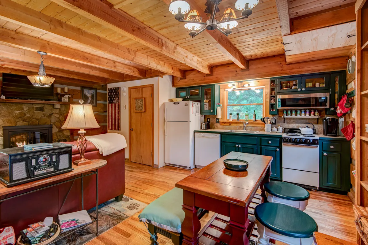 This image presents an expansive view of the living area, with impressive wooden beams above, a prominent stone fireplace, a cozy kitchen corner, and a dining table. The scene exudes rustic mountain charm, blending elements for gatherings and relaxation, and highlights the open layout that seamlessly connects each area.