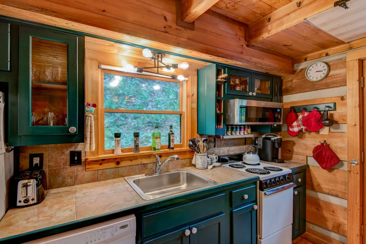 A warmly lit kitchen featuring green cabinetry, a window offering views of the surrounding landscape, modern appliances, and rustic decor elements. The butcherblock countertop and upscale amenities are clearly visible, evoking the essence of a mountain cabin kitchen.
