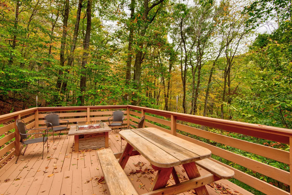 An inviting exterior space with a wooden picnic table, fire pit, comfortable chairs, and a surrounding forest. The area is set for outdoor dining and cozy gatherings, with the fire pit suggesting warmth and opportunities for connection after sunset.