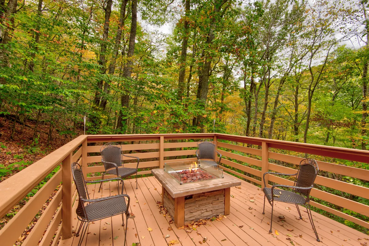 A cozy outdoor setup featuring a modern gas firetable surrounded by comfortable chairs, set against a backdrop of lush trees and the deck railing. The environment encourages gathering and relaxation while enjoying the wooded views.